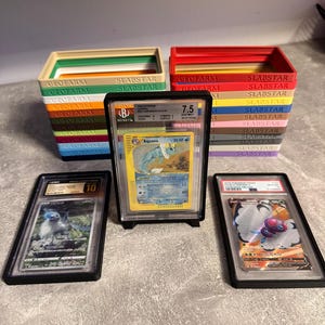 Pu&ograve; includere: Un assortimento di carte Pok&eacute;mon classificate in custodie protettive, esposte con contenitori colorati. Le carte presentano vari personaggi Pok&eacute;mon e sono classificate con valori numerici. I contenitori sono etichettati con il testo "GEOFARNI" e "SLABSTAR".