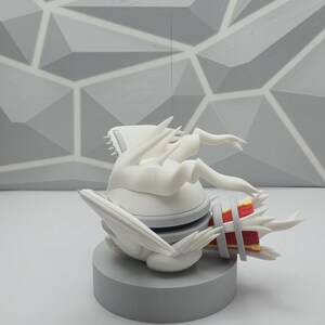 May include: A white, stylized creature figurine with sharp, wing-like protrusions and a red and yellow cylindrical tail section. The figure is mounted on a solid, circular gray base, featuring a smooth matte finish and intricate, angular details.
