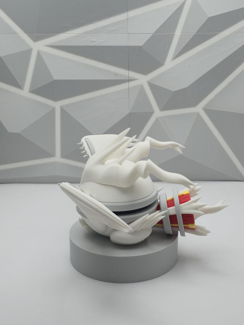 May include: A white, stylized creature figurine with sharp, wing-like protrusions and a red and yellow cylindrical tail section. The figure is mounted on a solid, circular gray base, featuring a smooth matte finish and intricate, angular details.