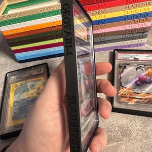 Pu&ograve; includere: Una custodia protettiva nera con la scritta "SLABSTAR" contiene una carta collezionabile valutata. Varie scatole di stoccaggio colorate con le etichette "GEOFARM" e "SLABSTAR" sono impilate sullo sfondo. Sono visibili anche altre due carte valutate.