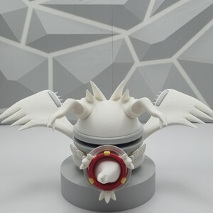 May include: A white 3D printed creature figurine featuring large wings, pointed head spikes, and a central circular red and yellow eye-like emblem. The figure is mounted on a solid gray cylindrical base, showcasing a smooth matte finish.