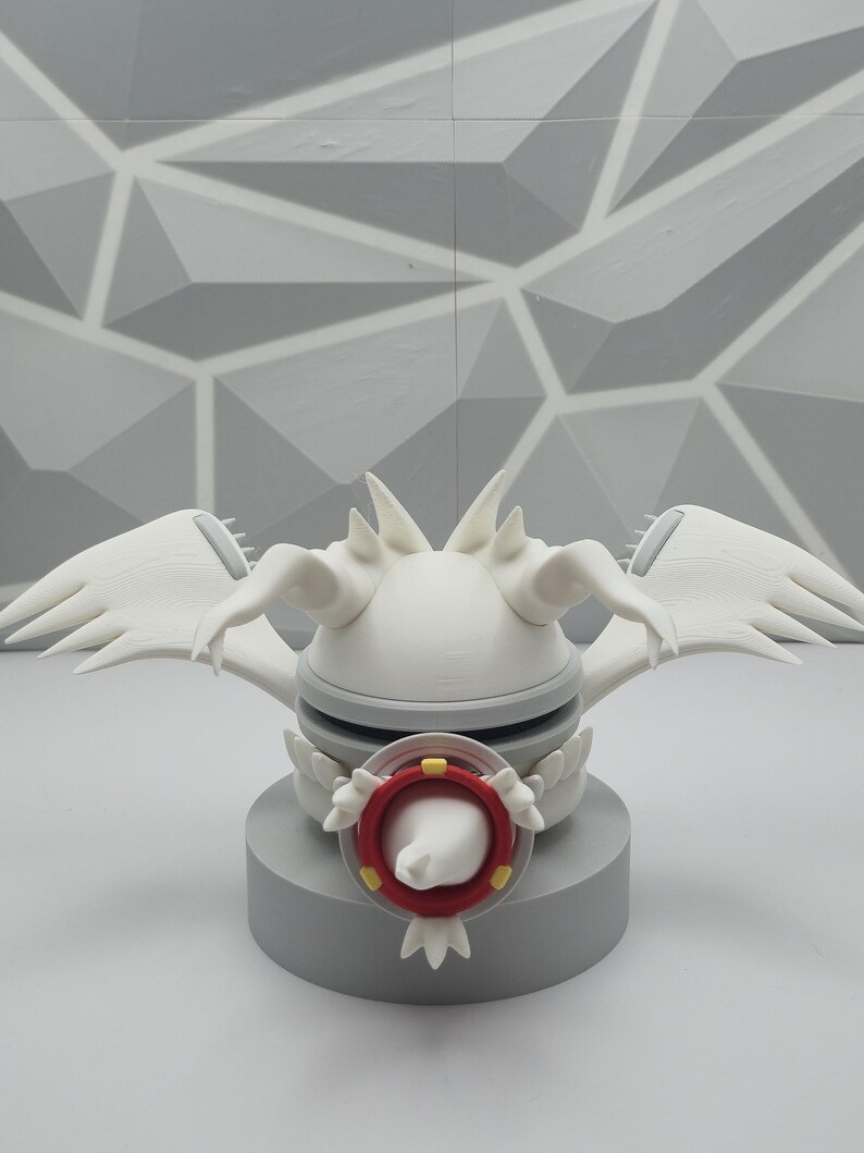 May include: A white 3D printed creature figurine featuring large wings, pointed head spikes, and a central circular red and yellow eye-like emblem. The figure is mounted on a solid gray cylindrical base, showcasing a smooth matte finish.