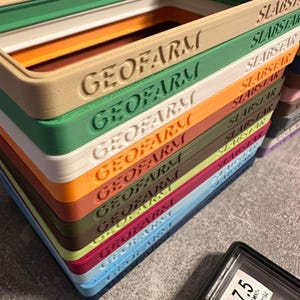 Pu&ograve; includere: Una pila di vassoi rettangolari colorati con il testo in rilievo "GEOFARM" e "SLABSTAR". I vassoi sono in vari colori, tra cui verde, beige, arancione e blu. Una carta valutata &egrave; visibile in basso a destra.