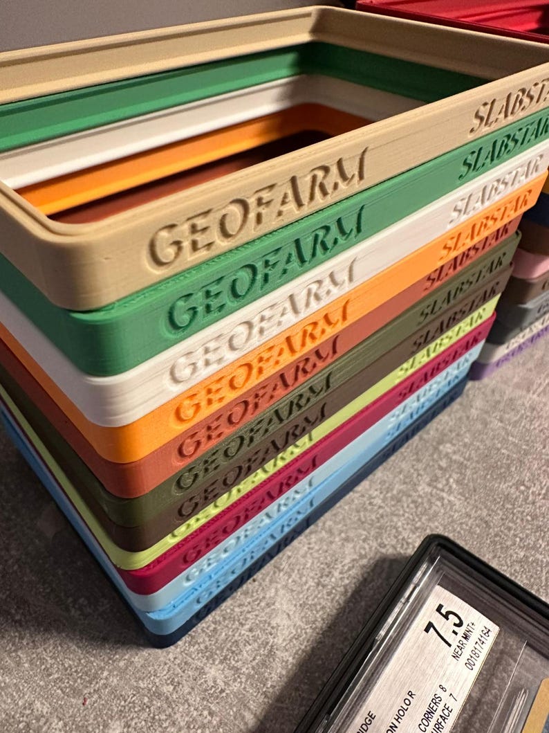 Pu&ograve; includere: Una pila di vassoi rettangolari colorati con il testo in rilievo "GEOFARM" e "SLABSTAR". I vassoi sono in vari colori, tra cui verde, beige, arancione e blu. Una carta valutata &egrave; visibile in basso a destra.