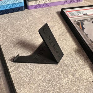 Pu&ograve; includere: Un espositore nero angolato con la scritta "SLABSTAND" stampata sul lato. Il supporto &egrave; progettato per contenere carte collezionabili. Pile di custodie per carte con le scritte "GEOFARM" e "SLABSTAR" sono visibili sullo sfondo.