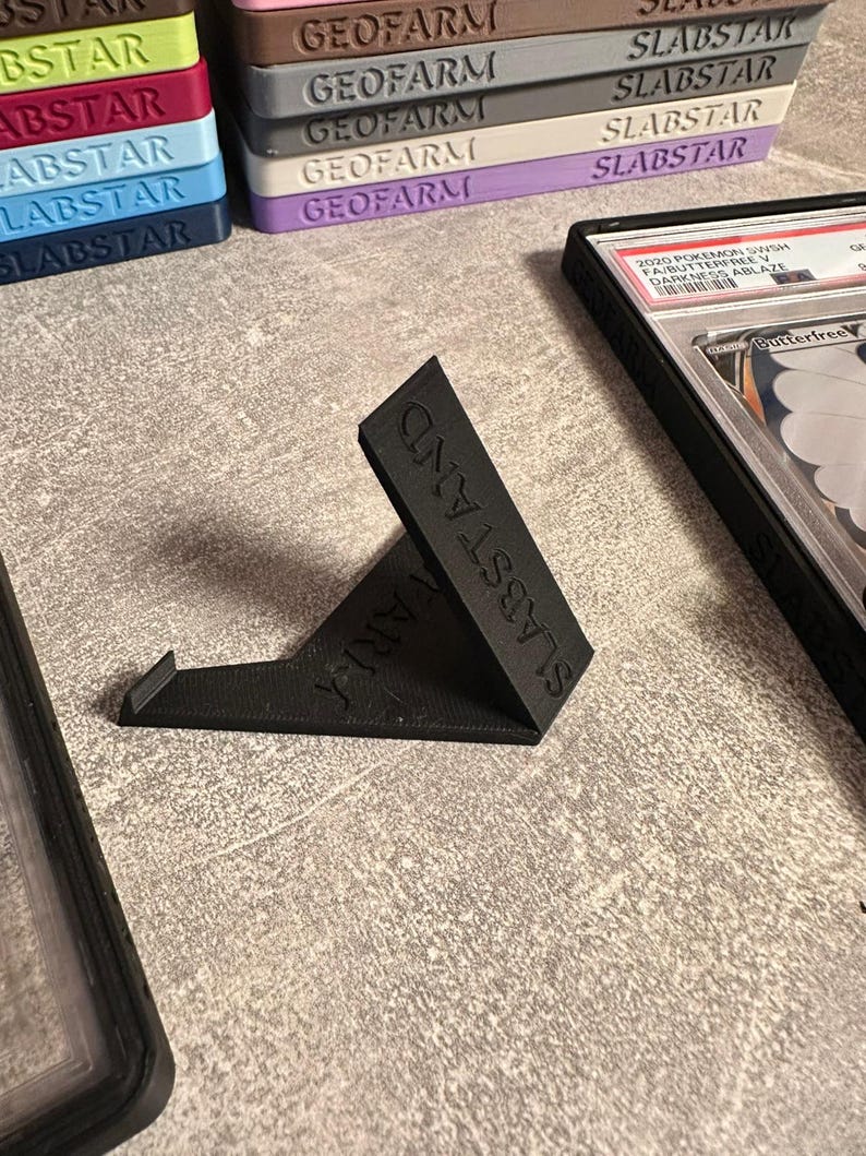 Pu&ograve; includere: Un espositore nero angolato con la scritta "SLABSTAND" stampata sul lato. Il supporto &egrave; progettato per contenere carte collezionabili. Pile di custodie per carte con le scritte "GEOFARM" e "SLABSTAR" sono visibili sullo sfondo.