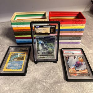 Pu&ograve; includere: Esposizione di carte Pok&eacute;mon classificate in custodie protettive, con pile di scatole colorate con etichetta "GEOFARM" e "SLABSTAR" sullo sfondo. Una carta &egrave; valutata 10 da CGC.