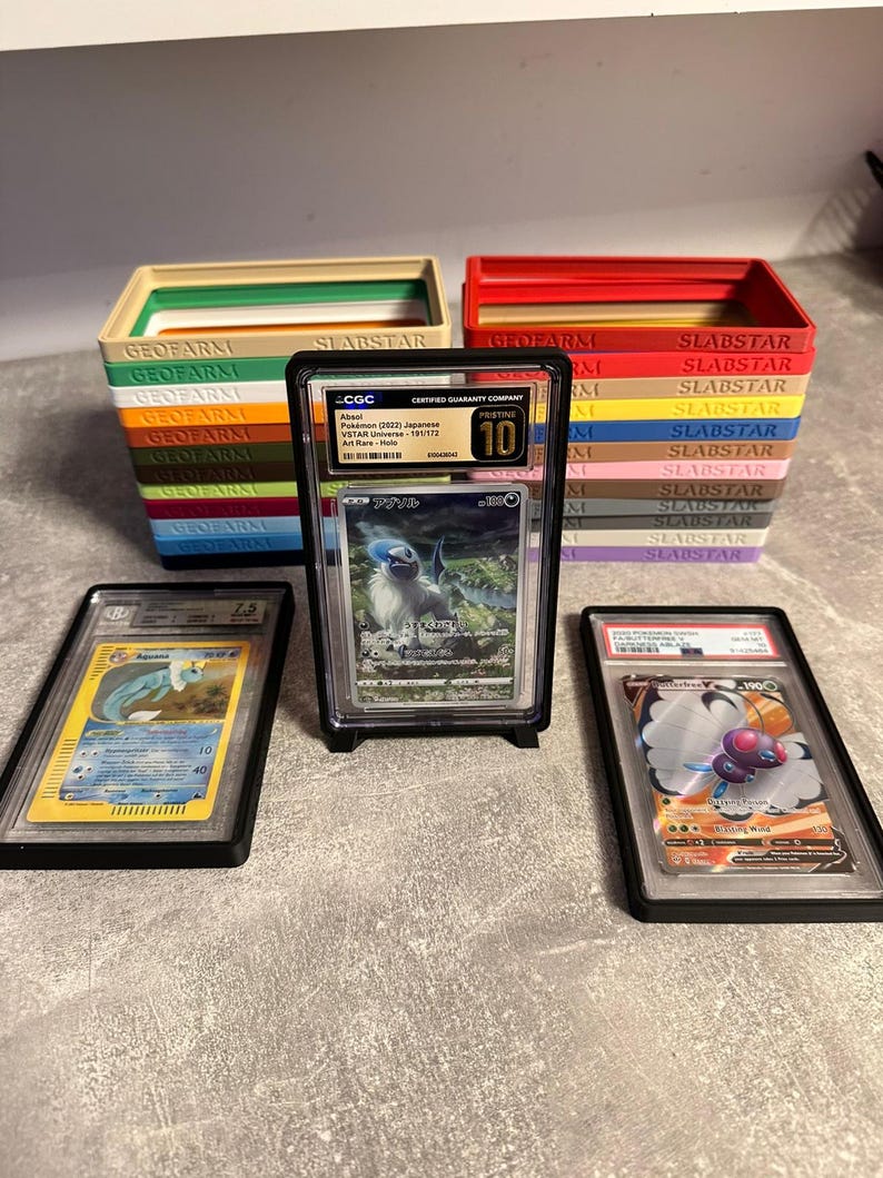 Pu&ograve; includere: Esposizione di carte Pok&eacute;mon classificate in custodie protettive, con pile di scatole colorate con etichetta "GEOFARM" e "SLABSTAR" sullo sfondo. Una carta &egrave; valutata 10 da CGC.