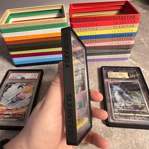 Pu&ograve; includere: Una pila di scatole colorate per la conservazione di carte con le scritte "GEOFARM" e "SLABSTAR". Sono visibili due custodie per carte classificate, una con una carta Pok&eacute;mon.
