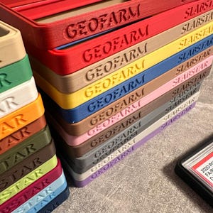Pu&ograve; includere: Una pila di cornici rettangolari colorate con i testi in rilievo "GEOFARM" e "SLABSTAR". Le cornici sono in vari colori, tra cui rosso, giallo, blu e rosa. Una custodia nera con testo bianco &egrave; visibile.