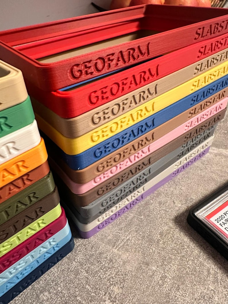 Pu&ograve; includere: Una pila di cornici rettangolari colorate con i testi in rilievo "GEOFARM" e "SLABSTAR". Le cornici sono in vari colori, tra cui rosso, giallo, blu e rosa. Una custodia nera con testo bianco &egrave; visibile.