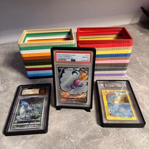 Pu&ograve; includere: Un assortimento di carte collezionabili Pok&eacute;mon classificate in custodie protettive. Le carte sono esposte davanti a pile di scatole colorate con la scritta "SLABSTAR" e "GEOFARM". Le carte presentano vari personaggi Pok&eacute;mon.