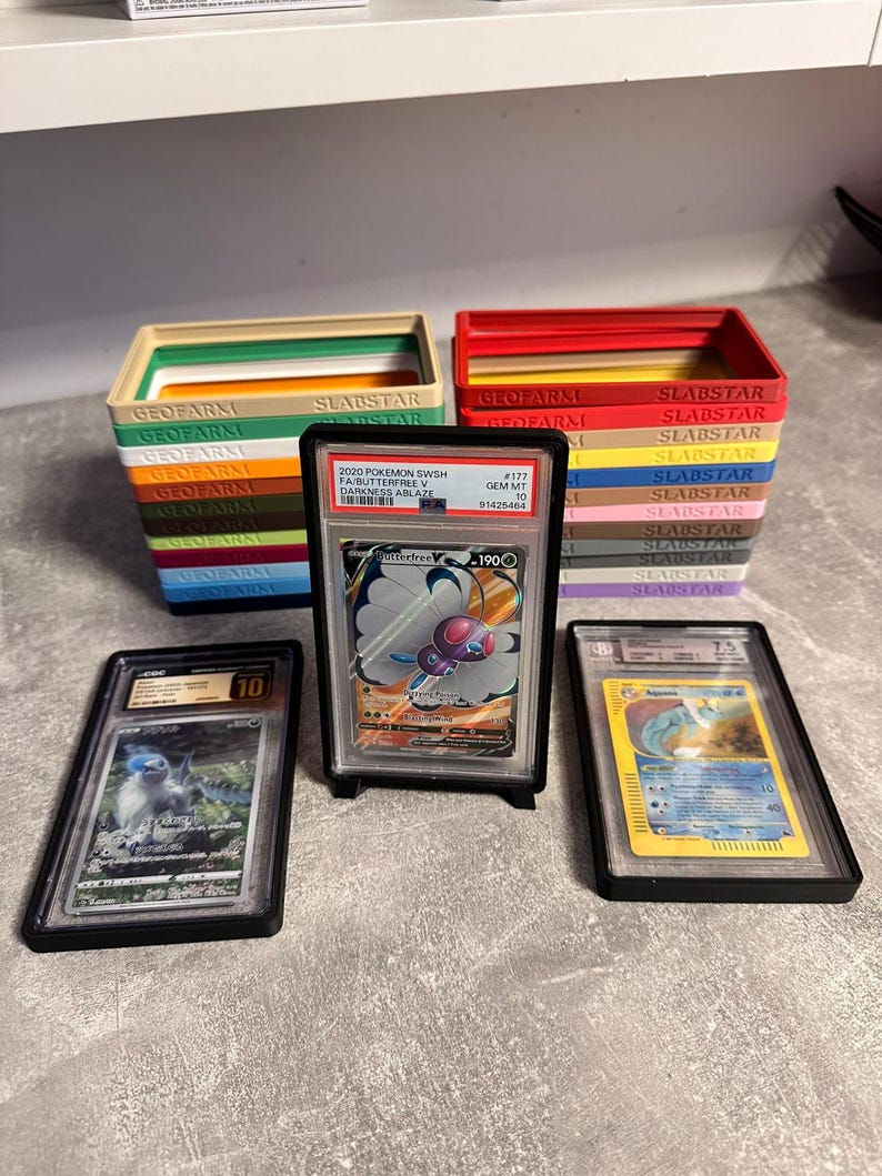 Pu&ograve; includere: Un assortimento di carte collezionabili Pok&eacute;mon classificate in custodie protettive. Le carte sono esposte davanti a pile di scatole colorate con la scritta "SLABSTAR" e "GEOFARM". Le carte presentano vari personaggi Pok&eacute;mon.