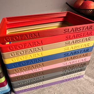 Pu&ograve; includere: Una pila di vassoi rettangolari colorati con la scritta "GEOFARM" e "SLABSTAR" stampata sopra. I vassoi sono di vari colori, tra cui rosso, giallo, blu e marrone. I vassoi sono impilati uno sopra l'altro.