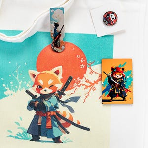 May include: A white canvas tote bag featuring a graphic of a red panda samurai with two swords against a large orange sun. The set includes a matching long bookmark with a samurai illustration and a small round pin depicting a panda face.