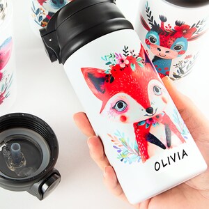 Puede incluir: Una colección de botellas de agua de acero inoxidable blanco con tapas negras abatibles. Cada botella presenta una colorida ilustración de un animal del bosque adornado con flores. Una botella muestra el nombre OLIVIA en texto negro.