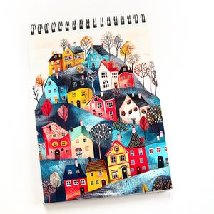 May include: Spiral-bound notebook with a vibrant illustration of a village on a hill. The image shows houses in shades of red, yellow, blue, and white, surrounded by trees and foliage. The notebook is set against a white backdrop.