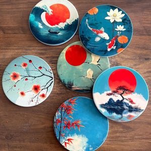 Op de afbeelding: Een set van zes ronde keramische onderzetters met diverse illustraties, waaronder een grote rode zon boven een golf, koivissen in een vijver, een bloeiende tak, vliegende kraanvogels, een bonsaiboom op een rotsachtig eiland en rode bamboebladeren.