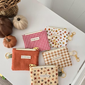 May include: A collection of small, zippered coin purses in various autumn-themed patterns and colors. Designs include red gingham, fall leaves, and solid orange and tan fabrics. Each purse has a gold-toned zipper and a small tag that reads "The Little Zinnia."