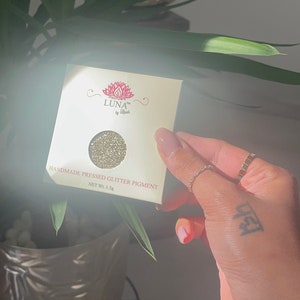 May include: A square, cream-coloured package of handmade pressed glitter pigment. The package features the brand name "LUNA by Rush" with a lotus flower logo. The glitter is a shimmering gold colour, and the net weight is 1.5g.