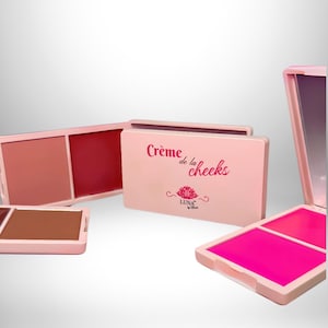 Op de afbeelding: Verschillende roze make-up compacts worden getoond, sommige open en andere gesloten. Een open compact toont twee blush tinten. Een gesloten compact heeft de tekst "Crème de la cheeks" en een lotusbloem logo. De make-up is van LUNA by J.Huan.