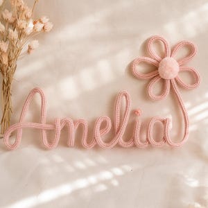 Pode incluir: Decoração de parede rosa tricotada com o nome "Amelia" em escrita cursiva, acompanhada por uma flor a condizer. A flor tem um centro de pompom e pétalas. Os artigos são colocados numa superfície clara.