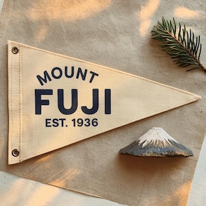 Può includere: Una bandiera a forma di pennone beige con le parole "MOUNT FUJI EST. 1936" in blu navy. Una piccola statuetta dipinta a mano del Monte Fuji con una cima innevata bianca è accanto alla bandiera. Un rametto di sempreverde è sullo sfondo.