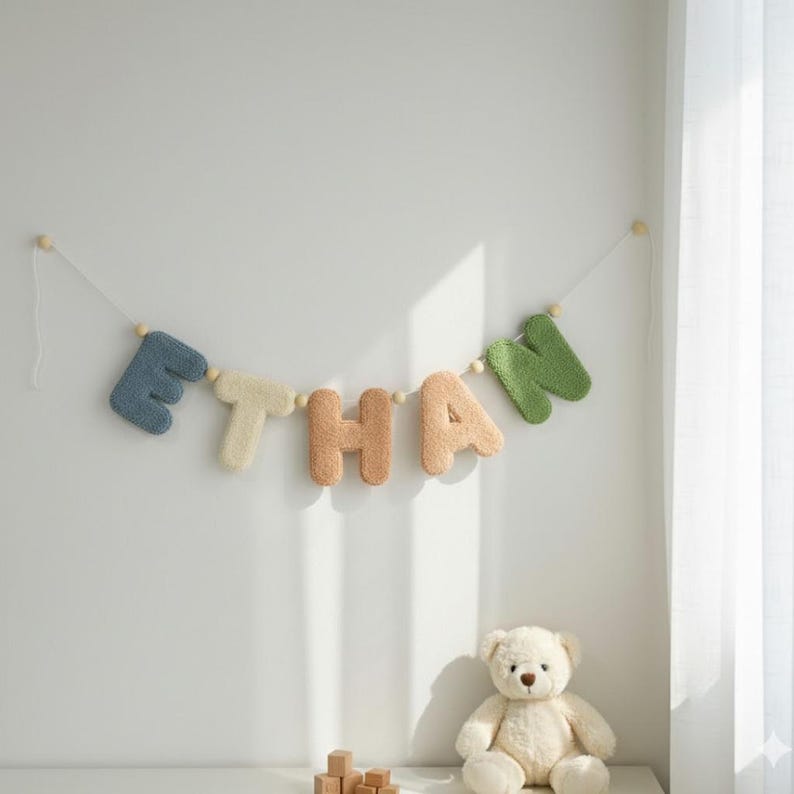 May include: A wall-mounted garland spelling the name ETHAN with individual plush, textured letters in blue, cream, tan, and green, separated by small wooden beads on a string, accompanied by a cream-colored plush teddy bear.