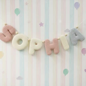 May include: A decorative garland spelling the name SOPHIA, featuring plush, textured letters in alternating shades of dusty pink, cream, and light gray, strung together on a thin cord with small wooden beads between each letter.