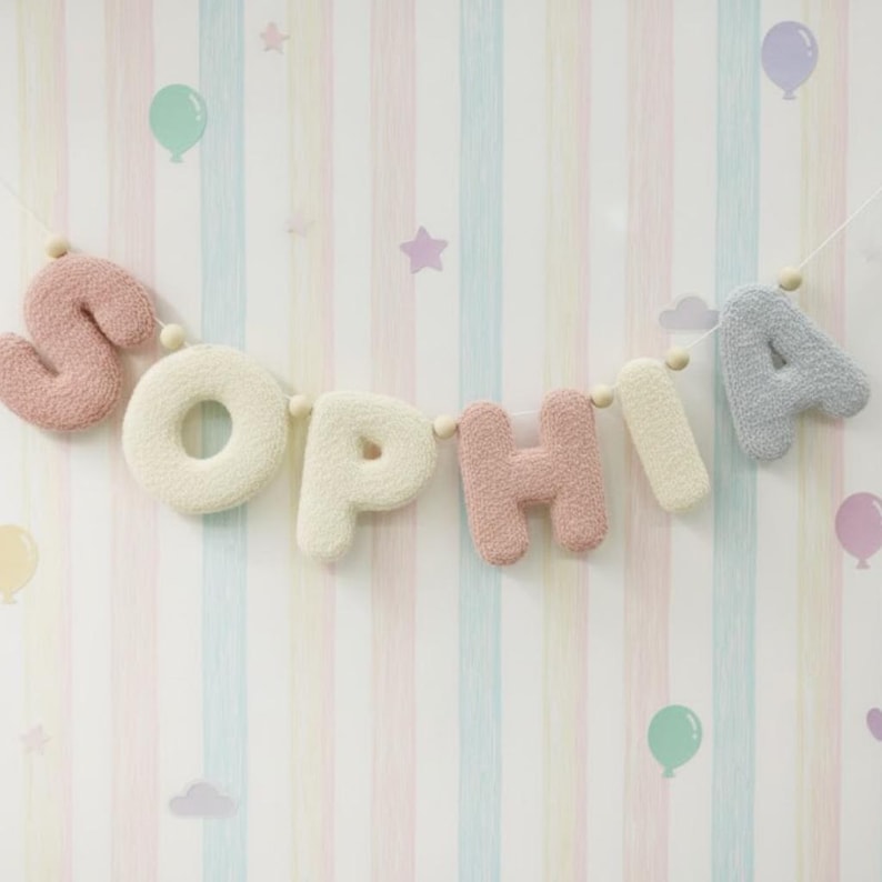 May include: A decorative garland spelling the name SOPHIA, featuring plush, textured letters in alternating shades of dusty pink, cream, and light gray, strung together on a thin cord with small wooden beads between each letter.
