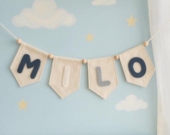May include: A decorative banner featuring four off-white fabric pennants strung together with wooden beads. The pennants display the name MILO in textured, raised letters, with the letters M and O in dark blue and I and L in light gray.