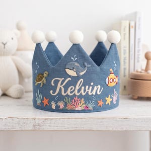 May include: A blue fabric crown featuring embroidered sea life including a whale, a sea turtle, a yellow submarine, and colourful coral. The name Kelvin is embroidered in white script in the centre. The crown is topped with five white pom-poms.