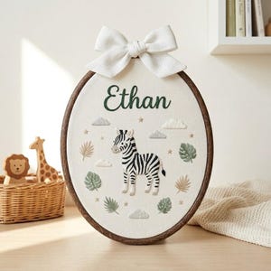 May include: An oval-shaped, embroidered wall hanging with a zebra design. The name "Ethan" is stitched in green above the zebra. The design includes clouds, stars, and leaves. A white bow is attached at the top. The frame is dark brown.
