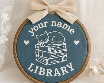 May include: A circular wooden embroidery hoop containing blue fabric with white stitched text that reads your name LIBRARY. The design features a white embroidered cat sleeping on a stack of three books, flanked by two small white embroidered hearts.