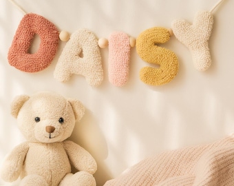 May include: A wall hanging banner spelling the name DAISY in tufted, textured letters of terracotta, cream, pale pink, and mustard yellow, separated by small wooden beads on a string, accompanied by a plush, light beige teddy bear.