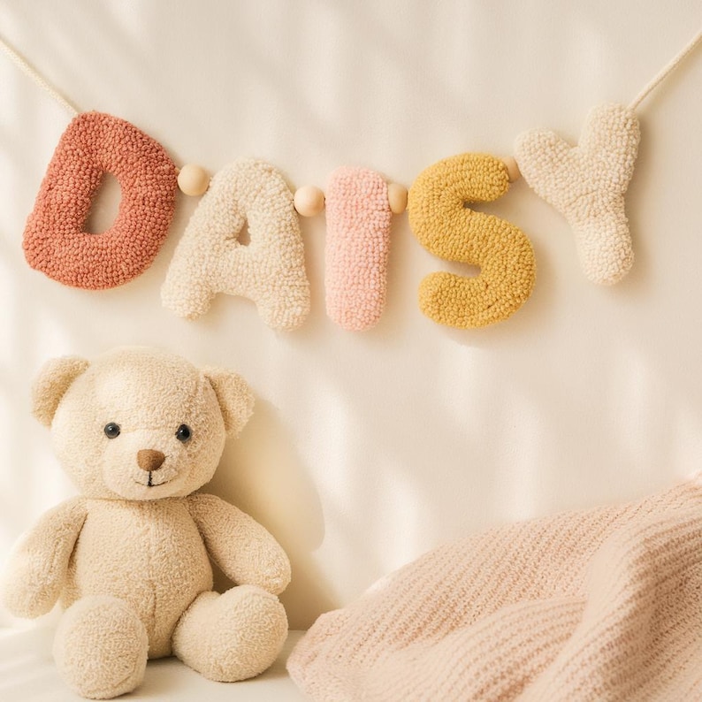 May include: A wall hanging banner spelling the name DAISY in tufted, textured letters of terracotta, cream, pale pink, and mustard yellow, separated by small wooden beads on a string, accompanied by a plush, light beige teddy bear.