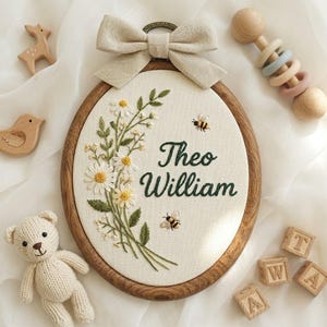 Op de afbeelding: Ovale borduurring met de naam "Theo William" in groene draad, omringd door geborduurde madeliefjes en bijen. Een beige strik siert de bovenkant. Houten speelgoed, waaronder een beer, een hert, een vogel en blokken, zijn eromheen geplaatst.