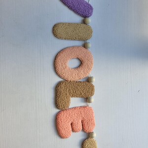May include: A vertical wall hanging featuring the word "VIOLET" spelled out in individual, tufted, textured fabric letters. The letters are colored in shades of purple, tan, peach, and light orange, separated by small, round, natural wooden beads.
