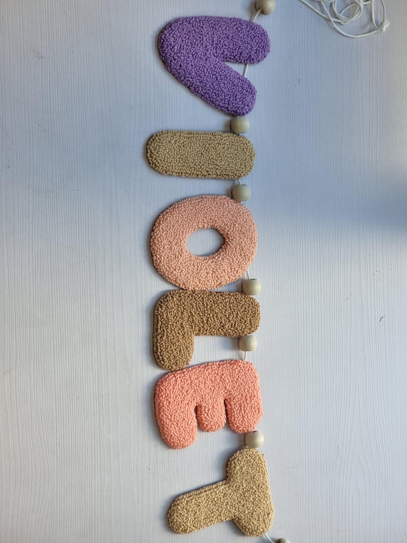 May include: A vertical wall hanging featuring the word "VIOLET" spelled out in individual, tufted, textured fabric letters. The letters are colored in shades of purple, tan, peach, and light orange, separated by small, round, natural wooden beads.