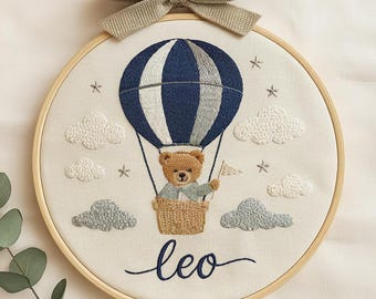 May include: Embroidered nursery art featuring a teddy bear in a hot air balloon with a navy blue and white striped design. The artwork includes embroidered clouds, stars, and the name "leo" in cursive. A beige bow is attached at the top.