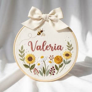 Op de afbeelding: Geborduurde hoepelkunst met de naam "Valeria" in sierlijke letters, omringd door zonnebloemen, madeliefjes en een bij. Een crèmekleurige strik is aan de bovenkant bevestigd. Het ontwerp is op crèmekleurige stof geplaatst.