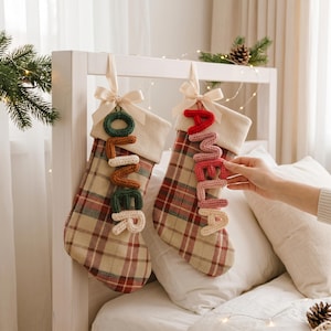 May include: Two Christmas stockings made of tan, red, and green plaid fabric with cream-colored cuffs and satin bows. One stocking features the name Oliver in stacked, knitted letters, and the other features the name Amelia in stacked, knitted letters.