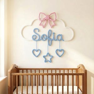 Puede incluir: Decoración de pared de guardería hecha a mano con el nombre "Sofia" en hilo azul, suspendida de un marco en forma de nube. Un lazo rosa se encuentra encima del nombre, con corazones azules y una estrella colgando debajo. La decoración está sobre una cuna de madera.