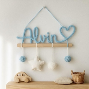 Puede incluir: Adorno de pared tejido en azul claro con el nombre "Alvin" y un corazón. Debajo, una barra de madera sostiene una estrella blanca, un pompón y dos bolas de hilo azules. Un coche de madera y una cesta de mimbre están en una estantería de madera.