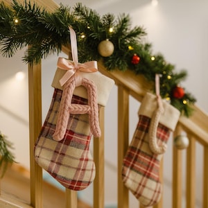 May include: Two Christmas stockings with red and beige plaid patterns hang from a wooden banister. Each stocking has a cream-colored cuff and a pink knitted letter or shape. A garland with lights and ornaments decorates the banister.