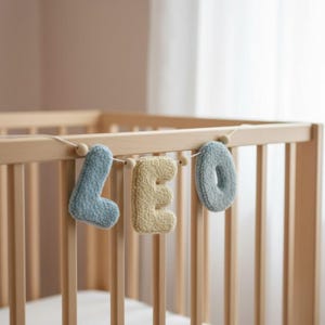 May include: A decorative garland featuring three plush, textured letters spelling LEO. The letters L and O are light blue, while the letter E is a soft yellow. Each letter is strung together on a thin cord with small wooden beads.