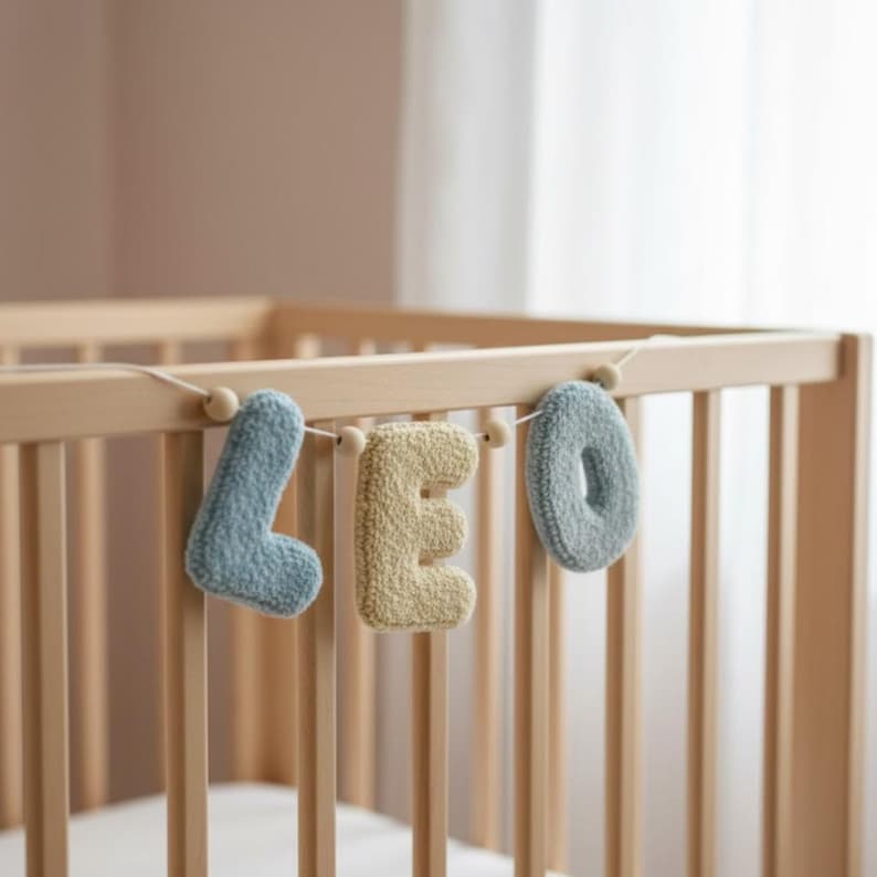May include: A decorative garland featuring three plush, textured letters spelling LEO. The letters L and O are light blue, while the letter E is a soft yellow. Each letter is strung together on a thin cord with small wooden beads.