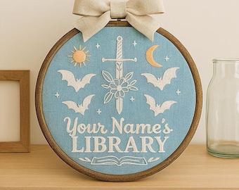 May include: A circular embroidery hoop featuring light blue fabric with white stitched designs of four bats, a sword, a flower, a sun, a crescent moon, and small stars. The text "Your Name's LIBRARY" is stitched in white above an open book.