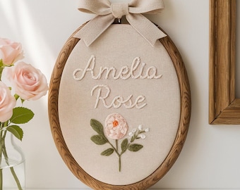 May include: An oval embroidery hoop featuring beige fabric with the name Amelia Rose stitched in white thread. Below the text is a small embroidered pink flower with green leaves and white buds. A beige fabric bow is attached to the top.