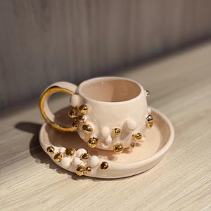 以下が含まれることがあります： クリーム色のセラミックティーカップとソーサーのセット。カップとソーサーは、金色の球状の装飾で飾られています。カップのハンドルと縁も金色のトリムです。セットは明るい色の木製の表面に表示されています。